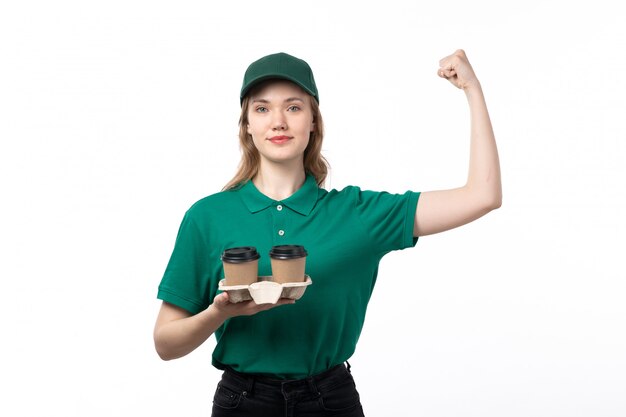 Una vista frontal joven mujer mensajero en uniforme verde con tazas de café flexionando y sonriendo en blanco