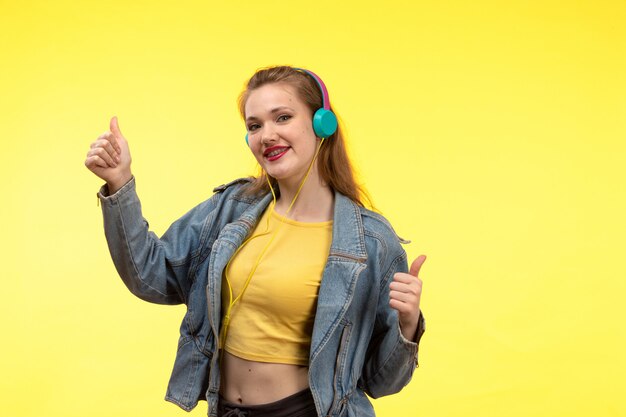 Una vista frontal joven mujer moderna en camisa amarilla pantalón negro y abrigo de jean con auriculares de colores escuchando música posando