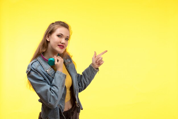 Una vista frontal joven mujer moderna en camisa amarilla pantalón negro y abrigo de jean con auriculares de colores posando sonriendo feliz