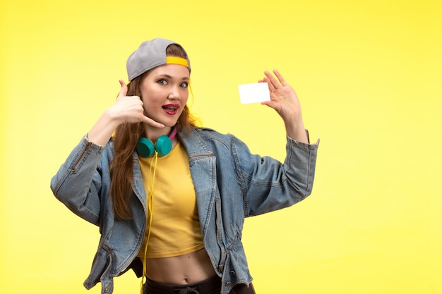 Una vista frontal joven mujer moderna en camisa amarilla pantalón negro y abrigo de jean con auriculares de colores con tarjeta blanca posando