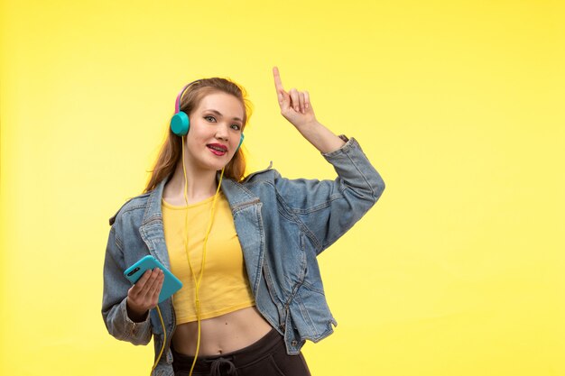 Una vista frontal joven mujer moderna en camisa amarilla pantalón negro y abrigo de jean con auriculares de colores con teléfono posando