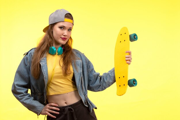 Una vista frontal joven mujer moderna en camisa amarilla pantalón negro y abrigo de jean sosteniendo patineta con auriculares de colores