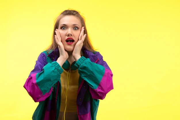 Una vista frontal joven mujer moderna en camisa amarilla pantalón negro y chaqueta colorida con auriculares de colores posando expresión sorprendida