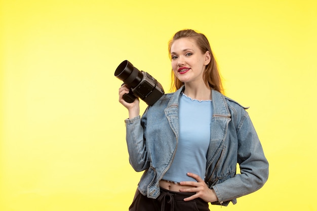 Una vista frontal joven mujer moderna en camisa azul pantalón negro y abrigo de jean posando feliz expresión sonriente sosteniendo la cámara de fotos