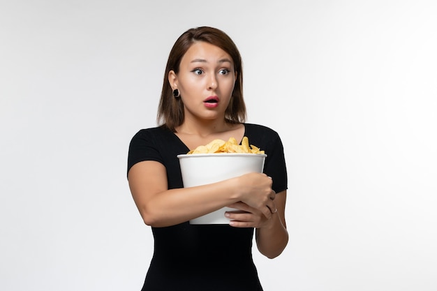 Vista frontal joven mujer sosteniendo la canasta con papas fritas y posando sobre una superficie blanca clara