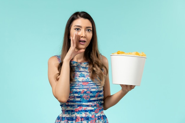 Vista frontal joven mujer sosteniendo la canasta con patatas fritas en la superficie azul claro