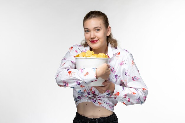 Vista frontal joven mujer sosteniendo la cesta con papas fritas y sonriendo sobre superficie blanca