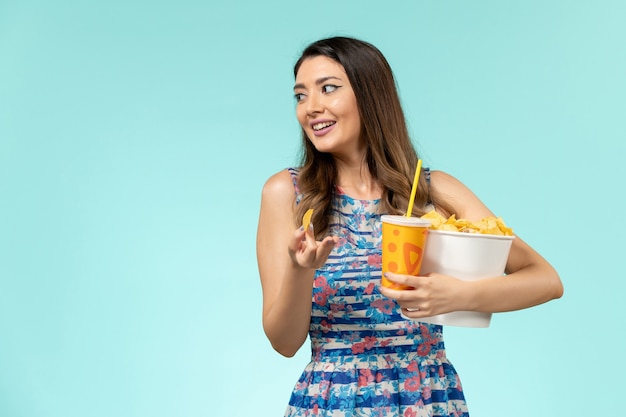 Vista frontal joven mujer sosteniendo la cesta con patatas fritas y bebida en la superficie azul