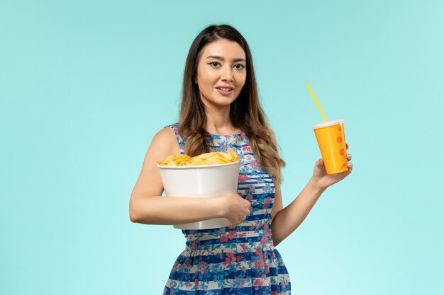 Vista frontal joven mujer sosteniendo la cesta con patatas fritas y bebida en la superficie azul