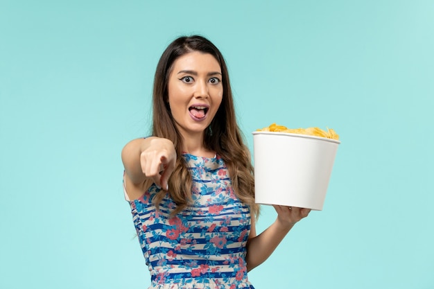 Vista frontal joven mujer sosteniendo la cesta con patatas fritas viendo la película y riendo sobre la superficie azul