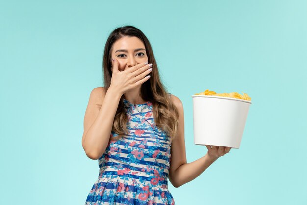 Vista frontal joven mujer sosteniendo la cesta con patatas fritas viendo la película y riendo sobre la superficie azul