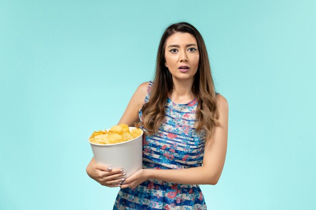 Vista frontal joven mujer sosteniendo la cesta con patatas fritas viendo la película en la superficie azul