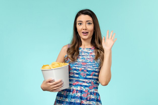 Vista frontal joven mujer sosteniendo la cesta con patatas fritas viendo la película en la superficie azul