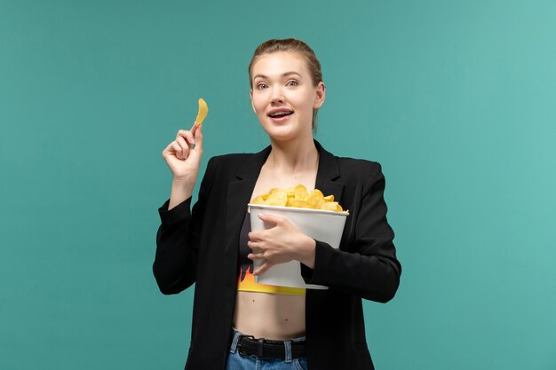 Vista frontal joven mujer sosteniendo y comiendo patatas fritas viendo la película en la superficie azul