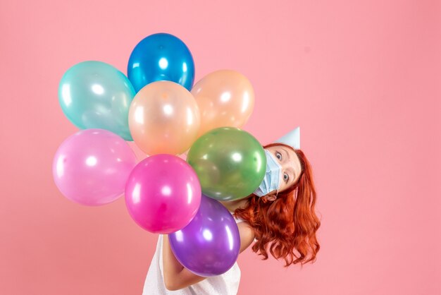 Vista frontal joven mujer sosteniendo globos de colores en máscara en rosa claro