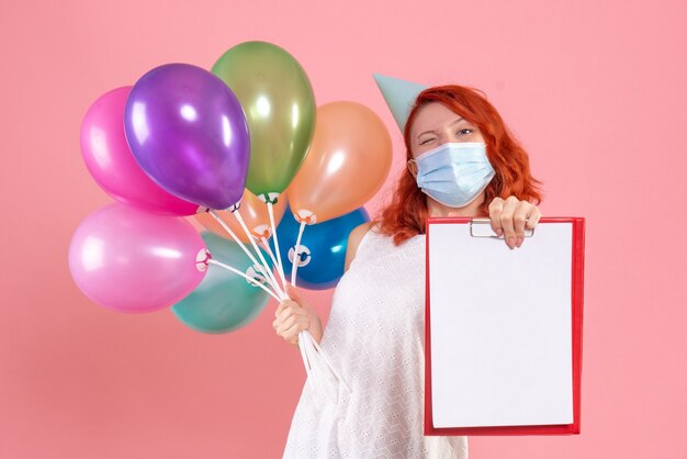 Vista frontal joven mujer sosteniendo globos de colores y nota de archivo en máscara en rosa