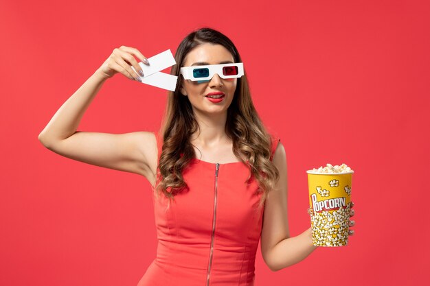 Vista frontal joven mujer sosteniendo palomitas de maíz con boletos en d gafas de sol en el escritorio rojo