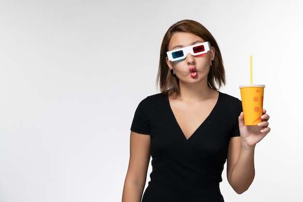 Vista frontal joven mujer sosteniendo refresco en d gafas de sol y haciendo muecas en la superficie blanca