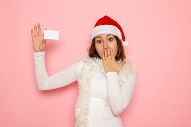 Vista frontal joven mujer sosteniendo una tarjeta bancaria blanca en la pared rosa Navidad color vacaciones año nuevo moda