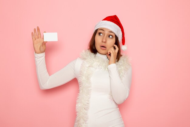 Vista frontal joven mujer sosteniendo una tarjeta bancaria blanca y pensando en la pared rosa Navidad vacaciones año nuevo moda color dinero