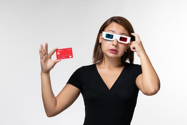 Vista frontal joven mujer sosteniendo una tarjeta bancaria en d gafas de sol y pensando en la superficie blanca