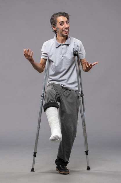 Foto gratuita vista frontal joven con muletas debido a un pie roto en un accidente de pared gris desactiva la pierna rota rota