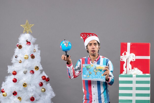 Vista frontal joven con ojos curiosos sosteniendo un mapa del mundo y un globo alrededor del árbol de Navidad y regalos
