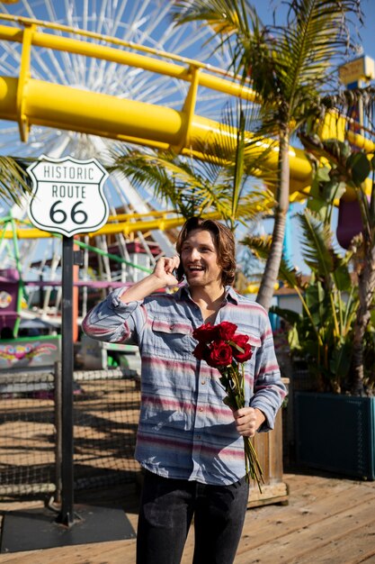 Vista frontal del joven en el parque de atracciones hablando por teléfono y sosteniendo un ramo de rosas