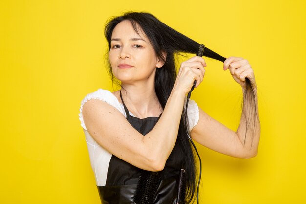 Una vista frontal joven peluquera en camiseta blanca capa negra con cepillos arreglando su cabello ondulado