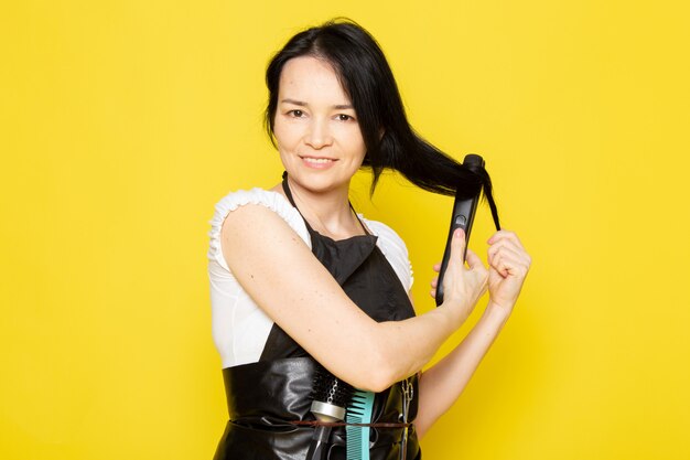 Una vista frontal joven peluquera en camiseta blanca capa negra fijación arreglando su cabello con herramienta sonriendo