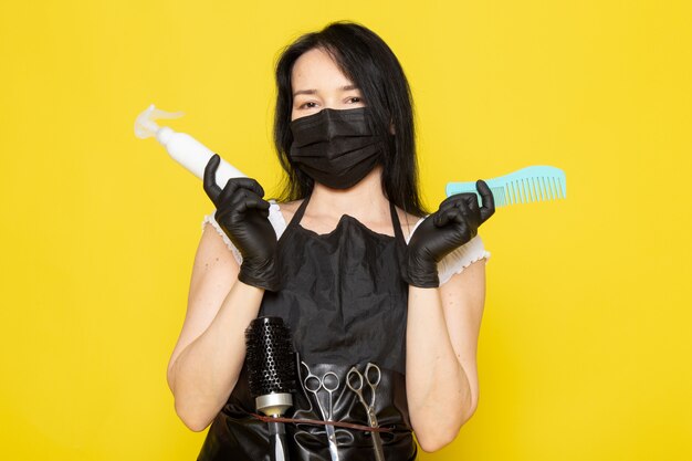 Una vista frontal joven peluquera en camiseta blanca capa negra con spray y cepillo para el cabello en máscara negra estéril guantes negros