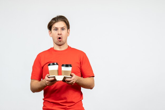 Vista frontal del joven preocupado en blusa roja sosteniendo café en vasos de papel sobre fondo blanco.