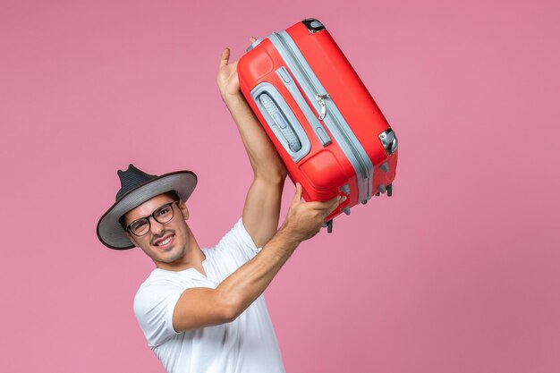 Vista frontal del joven que va de vacaciones y que sostiene la bolsa roja en la pared rosa