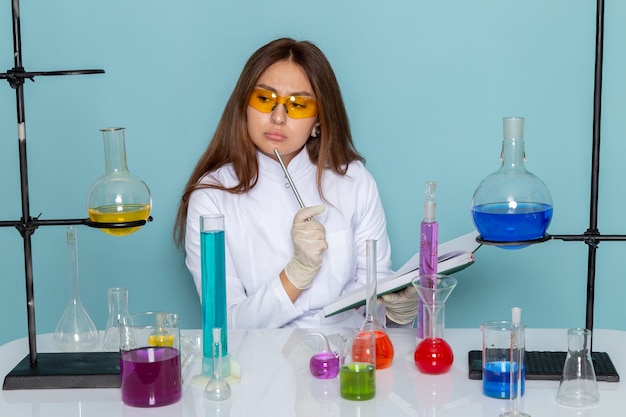 Vista frontal de la joven químico femenina en traje blanco delante de la mesa trabajando con soluciones y escribiendo notas