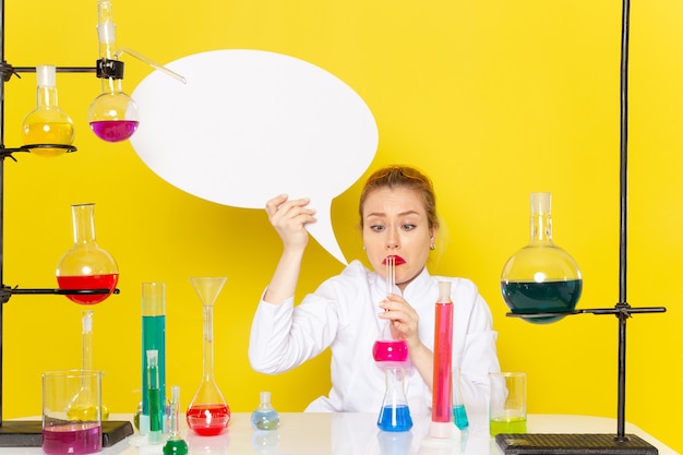 Foto gratuita vista frontal joven químico femenino sentado en traje blanco con diferentes soluciones con cartel blanco en la ciencia química espacial amarilla