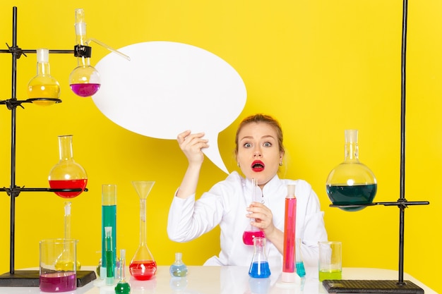 Foto gratuita vista frontal joven químico femenino sentado en traje blanco con diferentes soluciones con cartel blanco en el piso amarillo trabajo de ciencia química