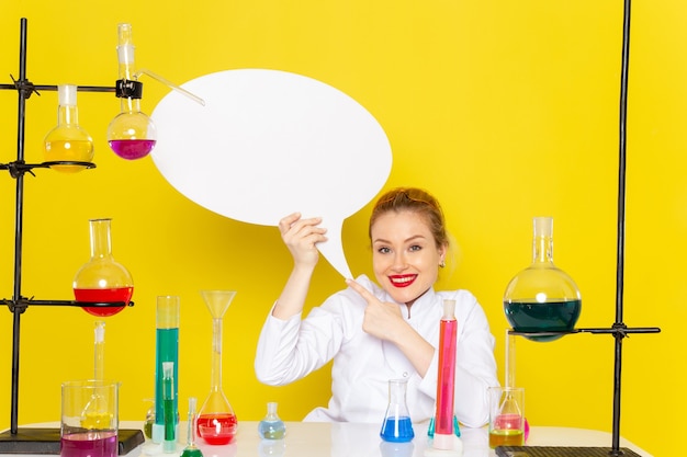 Foto gratuita vista frontal joven químico femenino sentado en traje blanco con diferentes soluciones con cartel blanco sonriendo en el piso amarillo ciencia química