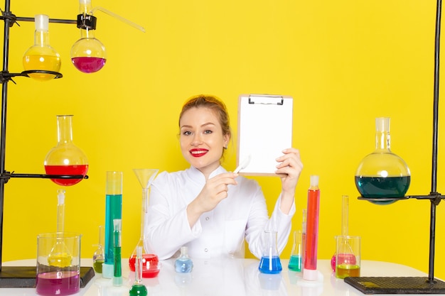 Vista frontal joven químico femenino sentado en traje blanco con diferentes soluciones sosteniendo el bloc de notas y sonriendo en el trabajo de proceso de ciencia de química espacial amarillo
