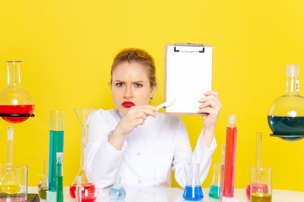 Vista frontal joven químico femenino sentado en traje blanco con diferentes soluciones sosteniendo el bloc de notas en el trabajo de proceso de ciencia de química espacial amarillo