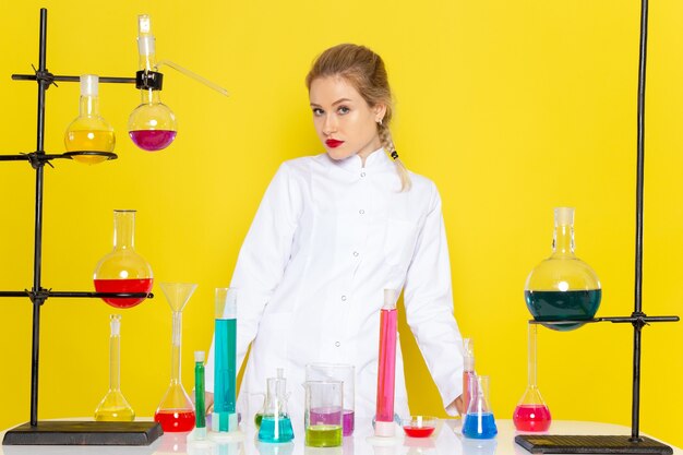 Vista frontal joven químico femenino en traje blanco frente a la mesa con soluciones ed en los experimentos científicos de química espacial amarillo