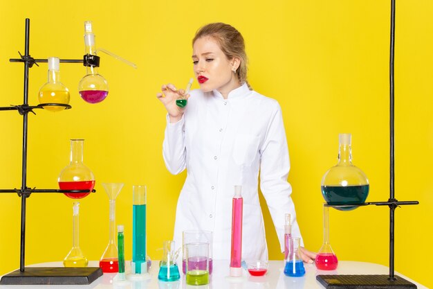 Vista frontal joven químico femenino en traje blanco frente a la mesa con soluciones ed sosteniendo uno en el experimento científico de química espacial amarillo