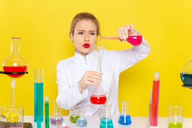 Vista frontal joven químico femenino en traje blanco frente a la mesa con soluciones ed trabajando con ellos mezclando la química espacial amarilla