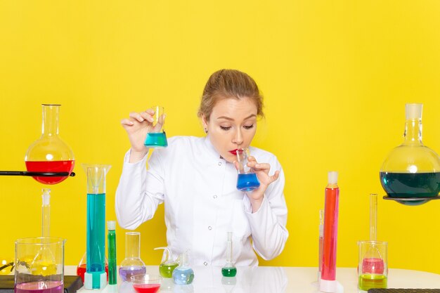 Vista frontal joven químico femenino en traje blanco frente a la mesa con soluciones ed trabajando con ellos en la química espacial amarilla