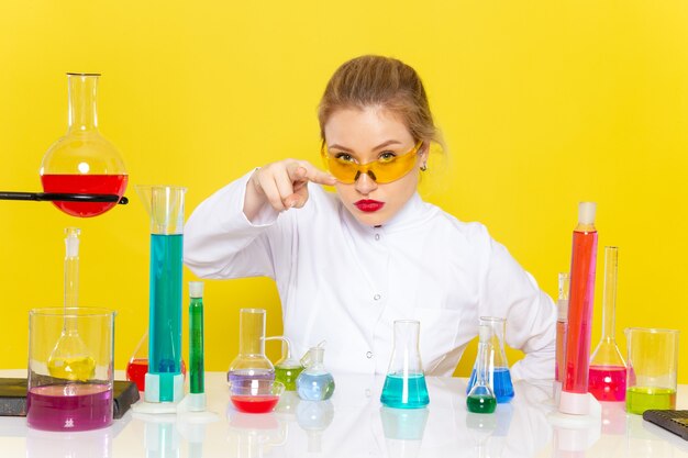 Vista frontal joven químico femenino en traje blanco frente a la mesa con soluciones ed trabajando con ellos sentados en la ciencia química espacial amarilla