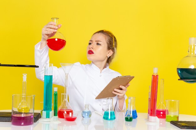 Vista frontal joven químico femenino en traje blanco con soluciones ed trabajando con ellos y sentado en el espacio amarillo ciencia química s