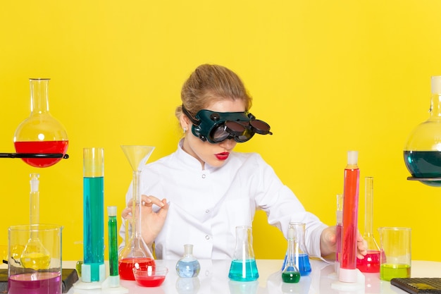Vista frontal joven químico femenino en traje blanco con soluciones ed trabajando con ellos sentado en máscara en el proceso de ciencia química espacial amarillo