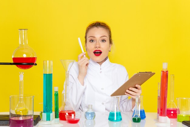 Vista frontal joven químico femenino en traje blanco con soluciones ed trabajando con ellos sosteniendo el bloc de notas en el espacio amarillo ciencia química s
