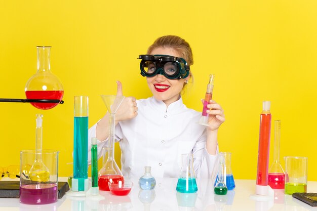 Vista frontal joven químico femenino en traje blanco con soluciones de educación trabajando con ellos y sentado con una sonrisa en la ciencia química espacial amarilla