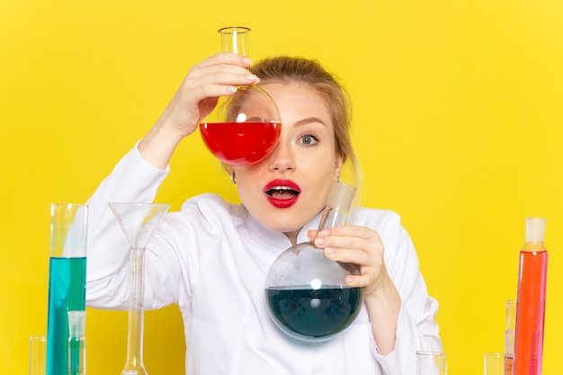 Vista frontal joven químico femenino en traje blanco sosteniendo diferentes soluciones en el trabajo de química espacial amarillo