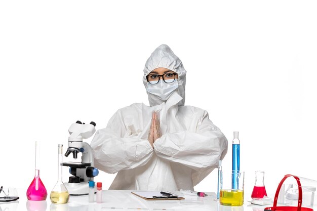 Vista frontal joven químico femenino en traje de protección especial sentado frente a la mesa con soluciones sobre fondo blanco laboratorio de virus covid salud química